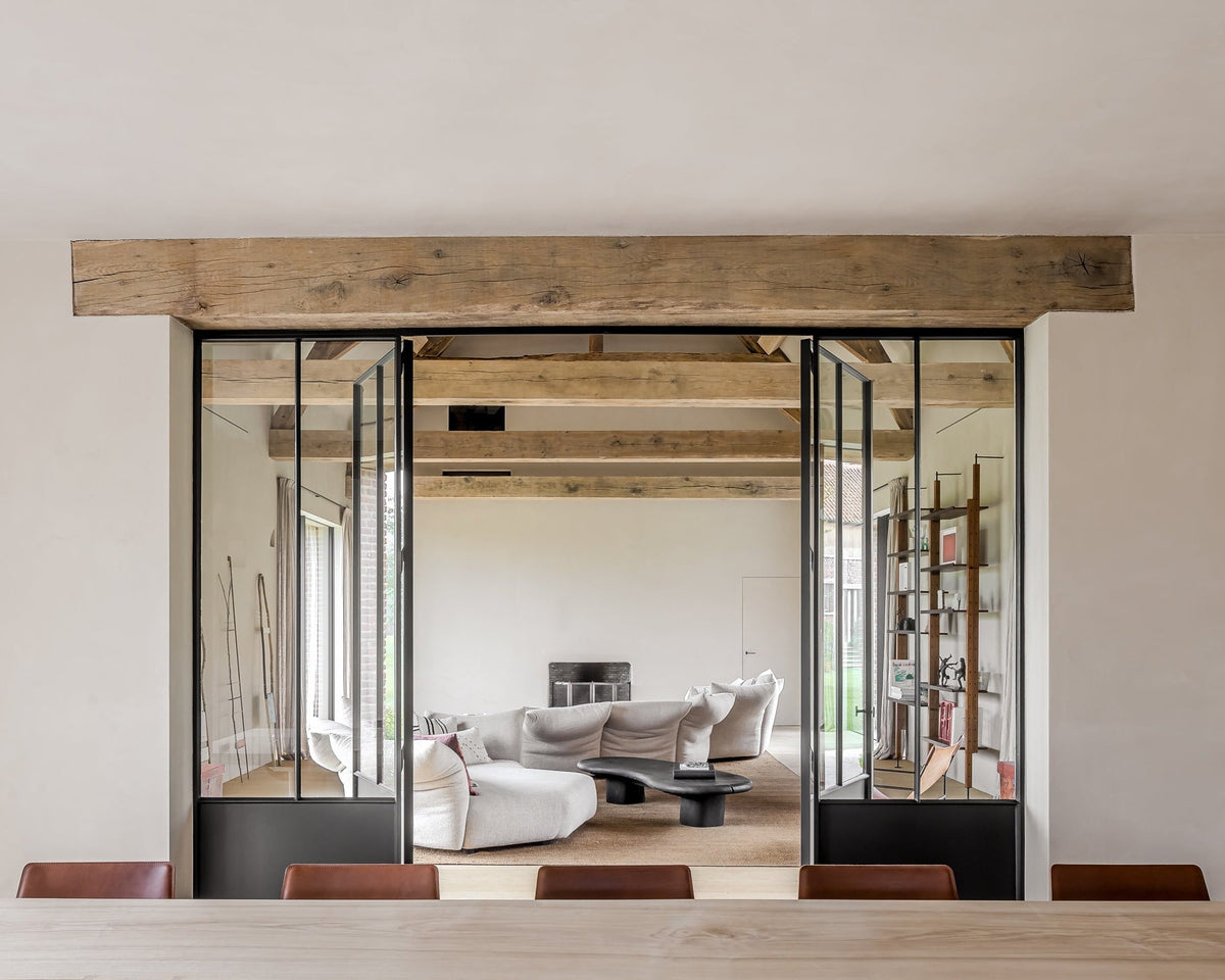 A modern interior space with large black-framed glass sliding doors, a rustic wooden ceiling beam, and a view into a living room with a light gray sofa and coffee table.